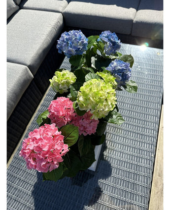 Die künstliche Hortensie Green UV 35 cm steht auf einem Korbtisch mit Glasplatte neben einem grauen Outdoor-Sofa mit realistischen grünen Hortensien, die im Sonnenlicht leuchten.