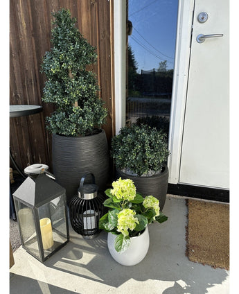 Auf der Veranda neben einer braunen Tür steht eine künstliche Hortensie Green UV 35 cm in einem weißen Topf mit gelbgrünen Blüten, daneben zwei Pflanzen in einem schwarzen Topf und zwei dekorative Laternen mit Kerzen.