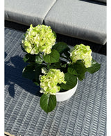 Die künstliche Hortensie Grün UV 35 cm mit hellgrünen Blüten und dunkelgrünen Blättern steht auf einem geflochtenen grauen Outdoor-Tisch mit einer gepolsterten Bank im Hintergrund.