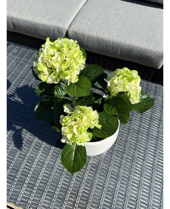 Die künstliche Hortensie Grün UV 35 cm mit hellgrünen Blüten und dunkelgrünen Blättern steht auf einem geflochtenen grauen Outdoor-Tisch mit einer gepolsterten Bank im Hintergrund.