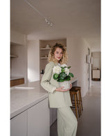 Eine Frau mit lockigen blonden Haaren, gekleidet in ein hellgrünes Outfit, steht in einer modernen Küche mit einer künstlichen Hydrangea White UV 35 cm Pflanze in ihren Händen – mit cremeweißen Blüten – und lächelt in die Kamera.