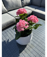 Die Kunstpflanze Hydrangea Pink UV 35 cm, ein weißer Topf mit leuchtend rosa Hortensienblüten, steht auf einem geflochtenen grauen Tisch im Freien mit einem grauen Sofa mit Kissen im Hintergrund, beleuchtet von Sonnenlicht und sanften Schatten.