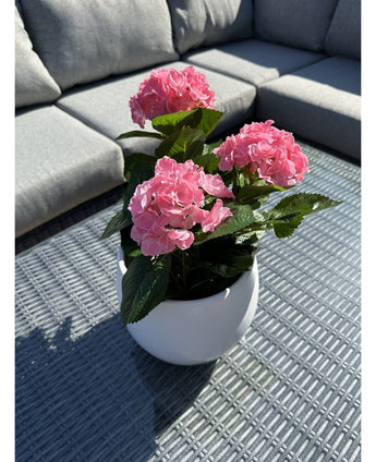 Die Kunstpflanze Hydrangea Pink UV 35 cm, ein weißer Topf mit leuchtend rosa Hortensienblüten, steht auf einem geflochtenen grauen Tisch im Freien mit einem grauen Sofa mit Kissen im Hintergrund, beleuchtet von Sonnenlicht und sanften Schatten.