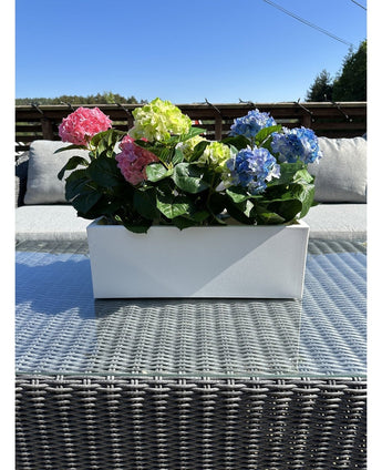 Die Kunstpflanze Hortensie Pink UV 35 cm, ein rechteckiger weißer Übertopf mit rosa Hortensien, steht draußen auf einem Korbtisch mit Glasplatte, grauen Kissen und einem blauen Himmel im Hintergrund.