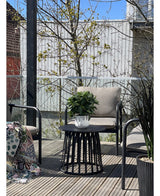 Eine moderne Terrasse mit zwei Stühlen mit Kissen, einem runden schwarzen Tisch mit der künstlichen Lorbeerpflanze UV ​​d33 cm als Mittelpunkt und einer gemusterten Decke auf einem Stuhl. Im Hintergrund erscheinen unter klarem Himmel Bäume und ein Holzzaun.