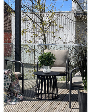 Eine moderne Terrasse mit zwei Stühlen mit Kissen, einem runden schwarzen Tisch mit der künstlichen Lorbeerpflanze UV ​​d33 cm als Mittelpunkt und einer gemusterten Decke auf einem Stuhl. Im Hintergrund erscheinen unter klarem Himmel Bäume und ein Holzzaun.
