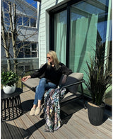 Eine Frau mit Sonnenbrille, schwarzem Top und blauen Jeans entspannt sich mit einem Drink in der Hand auf einem gepolsterten Stuhl auf einem sonnigen Balkon. Der Raum ist mit der künstlichen Lorbeer-UV-Pflanze d33 cm, einer lebendigen Dekoration, einer bunten Decke und einem kleinen Tisch mit Pflanzen ausgestattet.