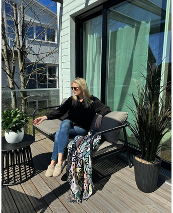 Eine Frau mit Sonnenbrille, schwarzem Top und blauen Jeans entspannt sich mit einem Drink in der Hand auf einem gepolsterten Stuhl auf einem sonnigen Balkon. Der Raum ist mit der künstlichen Lorbeer-UV-Pflanze d33 cm, einer lebendigen Dekoration, einer bunten Decke und einem kleinen Tisch mit Pflanzen ausgestattet.