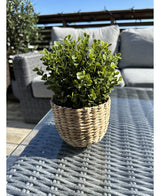 Die Kunstpflanze Buxus grün im UV-Topf 19 cm mit grünen Blättern in einem weidenkorbförmigen Topf steht auf einem Glastisch im Freien mit Terrassenmöbeln und einem Holzgeländer, das unter einem klaren blauen Himmel sichtbar ist.