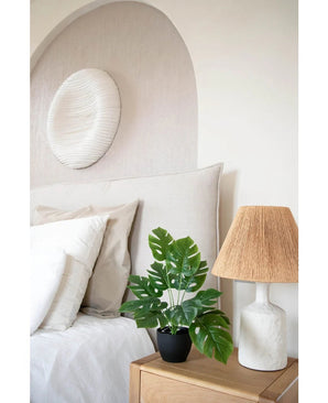 Ein modernes Schlafzimmer in neutralen Farbtönen verfügt über ein weißes Bett mit beigen und weißen Kissen, einen Nachttisch aus Holz mit einer Keramiklampe und einem gewebten Schirm, abstrakte Wandkunst über dem Bett und die Art Monstera 40 cm in einem schwarzen Dekotopf, der für viel Grün sorgt.