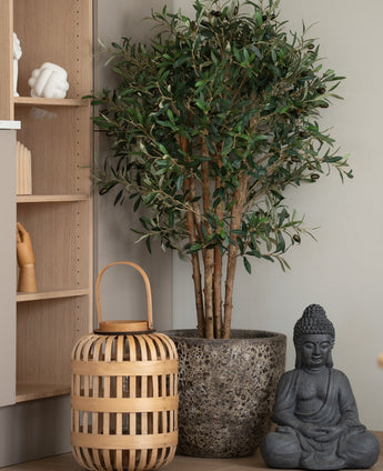 Ein künstlicher Olivenbaum UV 120 cm steht neben einer grauen Buddha-Statue und einer Holzlaterne auf dem Boden, daneben ein helles Holzbrett mit handgeformten Ornamenten. Diese pflegeleichte Pflanze bringt eine ruhige, gelassene Atmosphäre in den Raum.