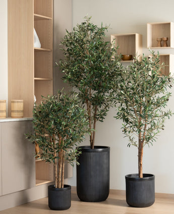 Der künstliche Olivenbaum UV 120 cm steht auf einem hellen Holzboden, im Hintergrund moderne helle Holzregale und Schränke. Der pflegeleichte, UV-beständige schwarze Rippentopf sorgt für einen modernen Look.