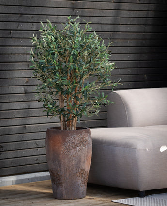 Der künstliche Olivenbaum UV 120 cm mit grünen Blättern steht neben einem hellgrauen Outdoor-Sofa auf einer Holzterrasse, im Hintergrund eine dunkle Holzlattenwand.