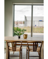 Ein Holztisch mit zwei Stühlen, ein künstlicher Palmensalon UV 60 cm in einem Topf und eine goldene Vase stehen neben einem großen Fenster mit Blick auf ein Industriegebiet mit Gebäuden und einem Zaun.