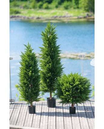 Drei künstliche Pflanzen-Koniferen-Deluxe-UV-120-cm-Bäume schmücken eine Holzterrasse mit Glasgeländer und Blick auf Wasser und Grün – dank ihres UV-Schutzes ideal für jeden Kunstpflanzengarten.