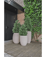 Drei große beige Pflanzgefäße mit künstlichen Conifer Deluxe UV 120 cm-Sträuchern stehen auf einer Holzterrasse in der Nähe einer schwarzen Tür und einer mit Efeu bedeckten Wand und bieten UV-Schutz für dauerhafte Schönheit in Ihrem Kunstpflanzengarten.