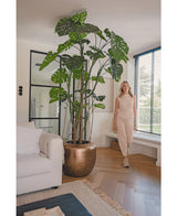 Eine Frau in beigem Outfit steht neben der künstlichen Monstera-Pflanze 240 cm in einem Bronzetopf. Das moderne Wohnzimmer verfügt über ein weißes Sofa, große Fenster mit Jalousien und einen Holzboden.