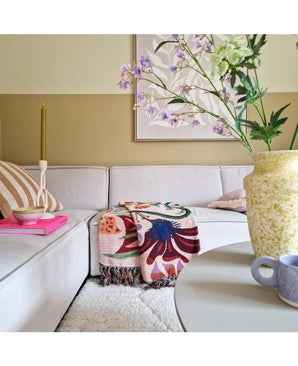 Ein gemütliches Wohnzimmer mit einem hellbeigen Sofa, einer bunten Decke mit Blumen und gemusterten Kissen. Auf einem runden Tisch steht eine gelbe Vase mit Kunsttak Daphne Spray 108 cm lila und eine kleine blaue Tasse. Kunst hängt an der neutralen Wand.