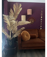 Ein modernes Wohnzimmer mit einem braunen Samtsofa, rundem Klappkissen und der Areca-Gold-Kunstpalme 120 cm in einer schwarzen Vase als Golddekor. Geometrische Wandkunst hängt an einer dunkelvioletten Wand, mit Blick auf Holzböden und vertikale Lamellen.