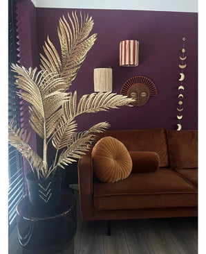 Ein modernes Wohnzimmer mit einem braunen Samtsofa, rundem Klappkissen und der Areca-Gold-Kunstpalme 120 cm in einer schwarzen Vase als Golddekor. Geometrische Wandkunst hängt an einer dunkelvioletten Wand, mit Blick auf Holzböden und vertikale Lamellen.