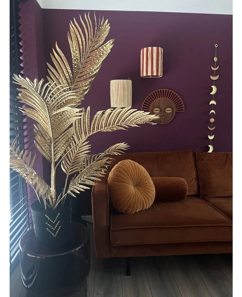 Ein modernes Wohnzimmer mit einem braunen Samtsofa, rundem Klappkissen und der Areca-Gold-Kunstpalme 120 cm in einer schwarzen Vase als Golddekor. Geometrische Wandkunst hängt an einer dunkelvioletten Wand, mit Blick auf Holzböden und vertikale Lamellen.
