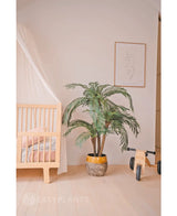 Een gezellige kinderkamer met een licht houten ledikant, de Kunstpalm Cycas Classic 135 cm in een gele plantenbak, een houten speelgoedfiets met het label MIA, minimalistische lijnkunst aan de muur, lichthouten vloeren en zachtroze beddengoed voor extra warmte.