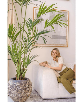 Eine Frau mit blonden Haaren, einem cremefarbenen Top und einer olivgrünen Hose sitzt lächelnd auf einem weißen Sofa und hält eine Tasse in der Hand. Die künstliche Kentia-Palme 220 cm verstärkt die tropische Atmosphäre des hellen, gemütlichen Raumes mit abstrakter Wandkunst.