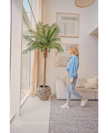 Eine Frau in blauer Bluse und Jeans geht an der Kunstpalm Phoenix De Luxe 210 cm vorbei an einem großen Fenster in einem hellen, modernen Wohnzimmer mit beigen Wänden, einem Sofa und gerahmter Kunst an der Wand.