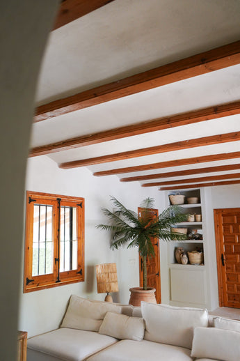 Ein gemütliches Wohnzimmer mit weißen Wänden, Holzbalken, einem weißen Sofagarnitur, der künstlichen Palme Phoenix De Luxe 210 cm, Holzfenster und -tür, eingebauten Regalen mit Körben und Töpferwaren und natürlichem Licht, das durch das Fenster fällt.