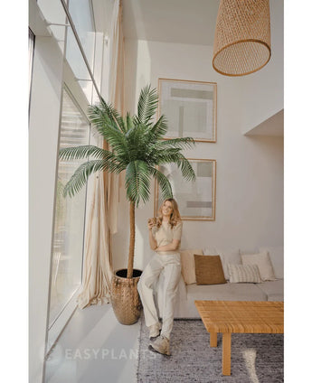 Eine Frau lächelt mit einem Drink in einem hellen, modernen Wohnzimmer, neben der Kunstpalme Phoenix De Luxe 210 cm. Der Raum verfügt über große Fenster, neutrales Dekor, gerahmte Kunst, ein Sofa, einen Holztisch und gewebte Akzente.