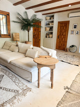 Ein gemütliches Wohnzimmer mit einem weißen Sofa, einem runden Beistelltisch aus Holz, einer künstlichen Palme Phoenix De Luxe 210 cm, geflochtenen Körben auf Regalen, Holztüren, einem runden Spiegel und Teppichen in neutralen Farbtönen auf einem hellen Boden.