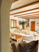 Ein gemütliches Wohnzimmer verfügt über ein großes cremefarbenes Sofa, einen Couchtisch aus Holz, eine künstliche Palme Phoenix De Luxe 210 cm in einem Topf, eine Holzbalkendecke, eingebaute Regale mit Keramik, natürliches Fensterlicht und eine warme, rustikale Einrichtung.