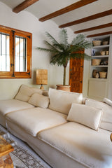 Ein gemütliches Wohnzimmer mit einem großen cremefarbenen Sofa, der Kunstpalme Phoenix De Luxe 210 cm in der Ecke, einer Holzlampe, eingebauten Regalen mit Körben, Holzdeckenbalken und einem Fenster mit Holzrahmen.