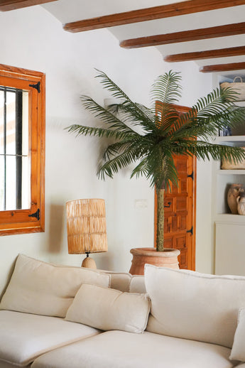 Ein gemütliches Wohnzimmer mit einem weißen Sofa, einer Rattan-Tischlampe, der Kunstpalme Phoenix De Luxe 210 cm im Topf, Holzbalken an der Decke, einem Fenster mit Holzrahmen und einer Holztür im Hintergrund.