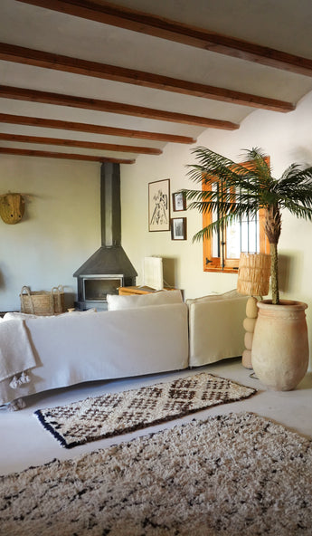 Ein gemütliches Wohnzimmer verfügt über ein weißes Sofa, einen gemusterten Teppich, einen Holzofen, gerahmte Kunstwerke und Holzbalken. Durch ein Fenster neben der üppigen Kunstpalme Phoenix De Luxe 210 cm strömt Sonnenlicht herein und erweckt den Raum zum Leben.