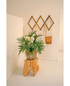 Die künstliche Pflanze Babypalmenfächer im Topf UV 50 cm steht auf einem Holzhocker in einem minimalistischen Raum. Dahinter präsentiert ein braunes Wandregal im Akkordeon-Stil braune und braune Ledertaschen vor einer strahlend weißen Wand.