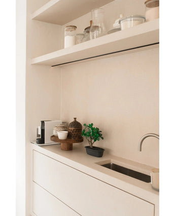 Minimalistische Arbeitsplatte mit einem kleinen Waschbecken, dekorativen Töpfen, einer Kaffeemaschine und Mülleimern auf schwebenden Regalen. In der neutralen Farbgebung Weiß-Beige wird der künstliche Bonsai Ficus 30 cm als Akzent-Topfpflanze verwendet.
