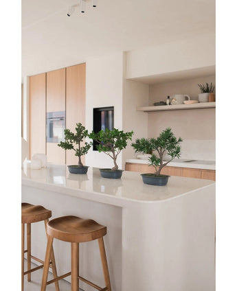 Eine künstliche Pflanze Bonsai Wacholder 40 cm steht auf einer weißen Kücheninsel mit zwei Holzhockern, umgeben von hellen Holzschränken, weißen Arbeitsplatten und einem minimalistischen Dekor.