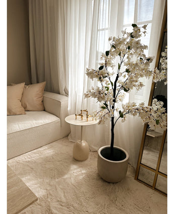 Eine gemütliche Wohnzimmerecke mit einem weißen Sofa, beigen Kissen, einem kleinen runden Beistelltisch mit goldenen Kerzenständern, transparenten Vorhängen und der künstlichen Bougainvillea 120 cm weiß für eine elegante Note.