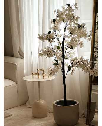 Eine künstliche Bougainvillea-Pflanze mit einer Größe von 120 cm steht neben einem goldumrandeten Spiegel neben einem kleinen runden weißen Beistelltisch mit goldenen Kerzenständern, hohen weißen Kerzen, transparenten weißen Vorhängen und einem cremefarbenen Sofa in einem hellen Raum.
