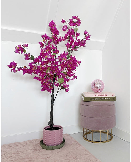 Die künstliche Bougainvillea 125 cm Lila mit leuchtend violetten Blüten steht neben einem rosa Samthocker mit darauf gestapelten Büchern und einer rosa Glaskugel in einem minimalistischen, modernen Raum mit weißen Wänden.