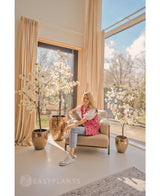 Eine Frau in einer rosa gemusterten Strickjacke und Jeans sitzt auf einem beigen Sessel an großen Fenstern mit cremefarbenen Vorhängen, umgeben von drei 180 cm großen weißen künstlichen Bougainvillea-Pflanzen in glänzenden goldenen Pflanzgefäßen. Sonnenlicht erfüllt den modernen, luftigen Raum.