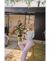 Eine blonde Frau in weißer Bluse und hellvioletter Hose sitzt draußen auf einem Hocker mit der künstlichen roten Topfpflanze Calathea Makoyana UV 75 cm auf einer Holzterrasse vor einem modernen Haus mit Glastüren.