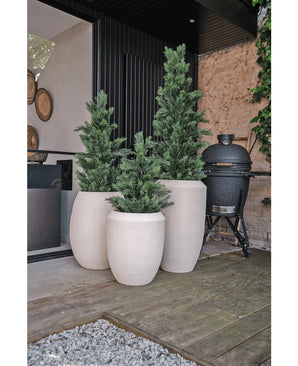 Auf einer Holzterrasse neben einem modernen Haus stehen drei hohe, weiße Pflanzgefäße mit Struktur und künstlichen Grünpflanzen „Conifer UV 120 cm“. In der Nähe steht ein schwarzer Außengrill mit einer Steinmauer und viel Grün im Hintergrund.