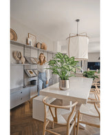 Eine Frau lächelt in einem hellen, modernen Esszimmer mit Holzstühlen, einem weißen Tisch mit der künstlichen Pflanze Dieffenbachia 75 cm als Mittelstück und offenen Regalen, die mit Büchern und Kunst dekoriert sind.
