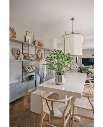Eine Frau lächelt in einem hellen, modernen Esszimmer mit Holzstühlen, einem weißen Tisch mit der künstlichen Pflanze Dieffenbachia 75 cm als Mittelstück und offenen Regalen, die mit Büchern und Kunst dekoriert sind.