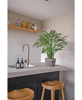 Eine moderne Küche mit einer Arbeitsplatte aus Beton, zwei Holzhockern und einer künstlichen Dieffenbachia-Pflanze (75 cm) neben dem Messinghahn, dazu Seifenspender und ein Holzregal mit Töpfen vor einer hellgrauen Wand.