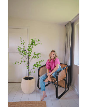 Eine Frau in einem rosa Top und blauen Jeans sitzt barfuß auf einem modernen Korbstuhl neben der künstlichen Pflanze Enkianthus 150 cm in einem hellen, minimalistischen Raum mit weißen Wänden und hellem Teppichboden.