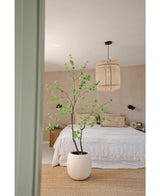 Ein minimalistisches Schlafzimmer mit der künstlichen Pflanze Enkianthus 180 cm als große Topfpflanze, einem Bett mit weißer Bettwäsche, einer gewebten Hängelampe, beigen Wänden und einem Naturfaserteppich für eine schlichte und luftige Atmosphäre.