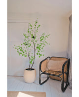 In einer minimalistischen Zimmerecke steht die Kunstpflanze Enkianthus 210 cm, eine blattgrüne Topfpflanze, neben einem Rattanstuhl mit schwarzem Gestell auf weißem Holzboden, mit neutralen Wänden und einem hellbeigen Vorhang.